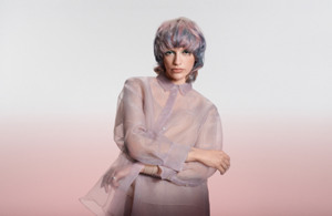 white woman with long blonde hair, which stands in front of a clean grey background looking into the camera